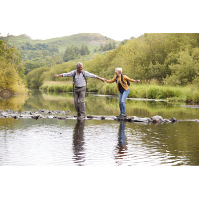 Millwood Pines Senior Couple Crossing River Whilst Hiking in UK Lake District by Monkeybusinessimages - Wrapped Canvas Photograph